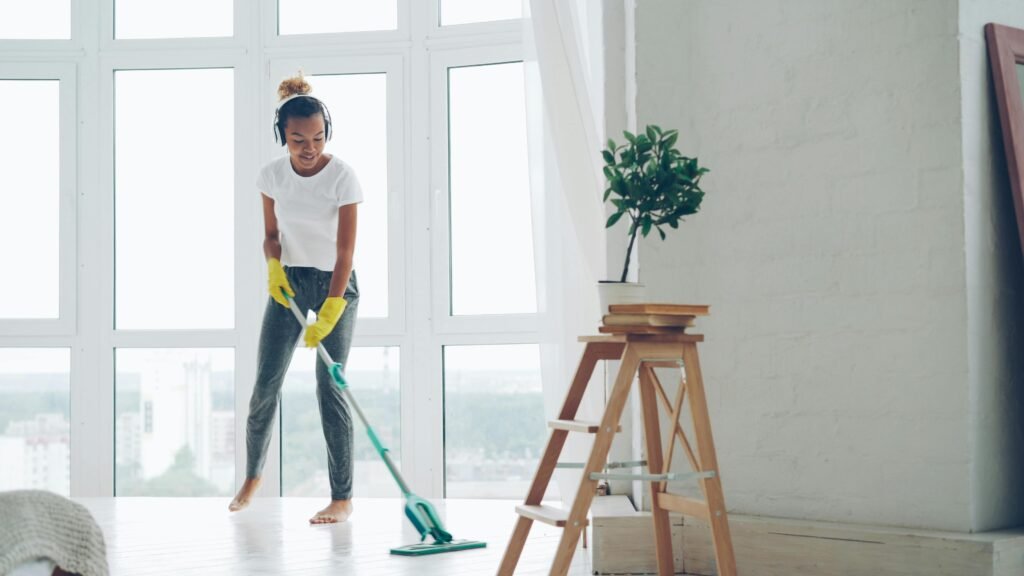 Young woman cleaning floor with a mop in a sunny room with large windows.