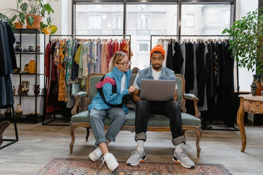 A couple sits on a couch in a stylish clothing store, browsing a laptop together.