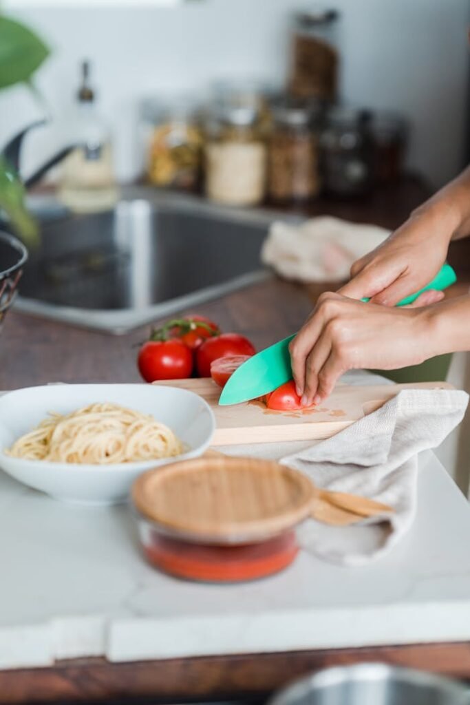 A person slices tomatoes with a green knife in a kitchen setting, alongside fresh pasta.